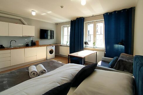A modern studio living area with a white kitchen along the left, a washing machine, a wall-mounted TV, and a wooden table by large windows with blue curtains.
