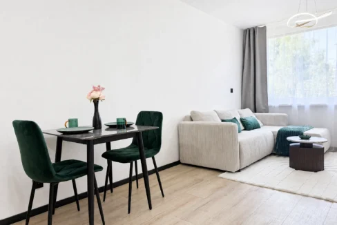Dining area with black table and green upholstered chairs, a vase of pink flowers, and a beige sofa in a bright room with large windows and sheer curtains.