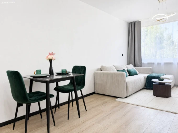 Dining area with black table and green upholstered chairs, a vase of pink flowers, and a beige sofa in a bright room with large windows and sheer curtains.